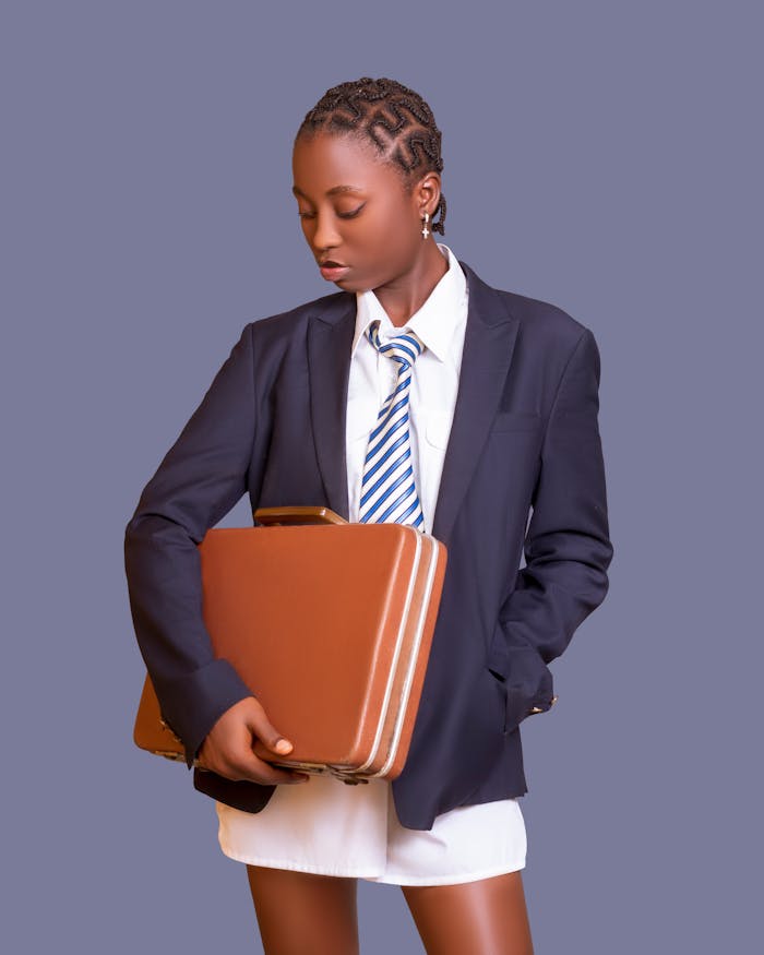 African American woman in a gray blazer holding a suitcase in a studio setting.