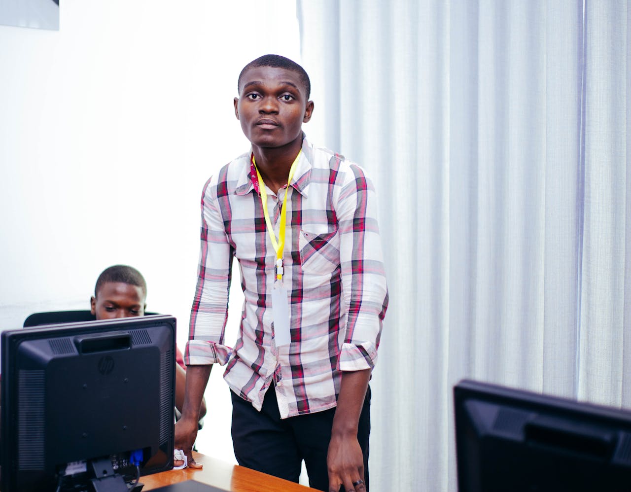 Young man in checked shirt engages in teaching session with computers in classroom.